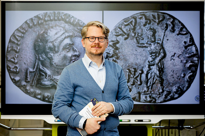 Gunnar Dumke has examined 4,500 coins. The image of a silver tetradrachm of the Indo-Greek ruler Archebios is on the monitor behind him (coin collection of the Berlin State Museums, photographer: Lutz-Jürgen Lübke).